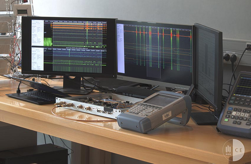 Stanowisko badawcze, widać trzy połączone monitory z uruchomionymi programami, podłączone urządzenie elektroniczne.